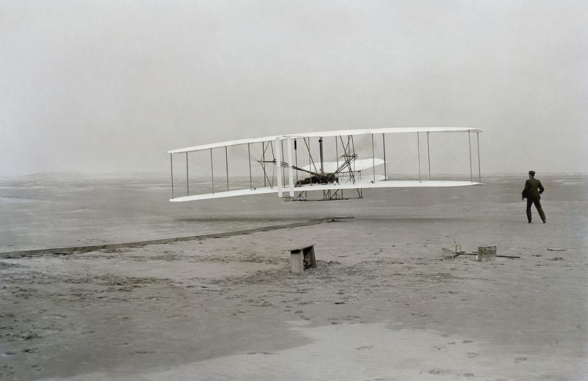 Picture taken of the first successful flight of the Wright Flyer. The machine traveled 120 ft (36.6 m) in 12 seconds at 10:35 a.m. at Kill Devil Hills, North Carolina. Orville Wright was at the controls of the machine, lying prone on the lower wing with his hips in the cradle which operated the wing-warping mechanism. Wilbur Wright ran alongside to balance the machine, and just released his hold on the forward upright of the right wing in the photo. The starting rail, the wing-rest, a coil box, and other items needed for flight preparation are visible behind the machine. This is described as "the first sustained and controlled heavier-than-air, powered flight" by the Fédération Aéronautique Internationale, but is not listed by the FAI as an official record. (Source: Wikipedia)